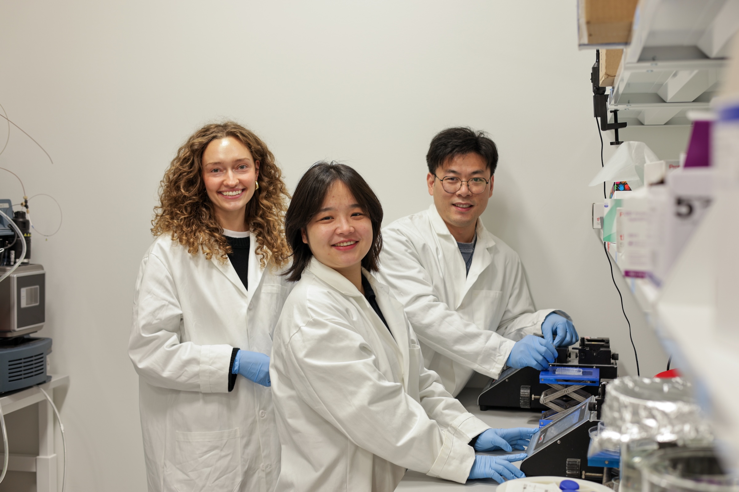 Three scientists in lab coats and rubber gloves demonstrate a piece of equipment on a lab bench.