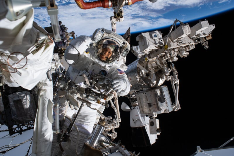 An astronaut in a spacesuit works outside the International Space Station, surrounded by equipment, with Earth visible in the background against the blackness of space.