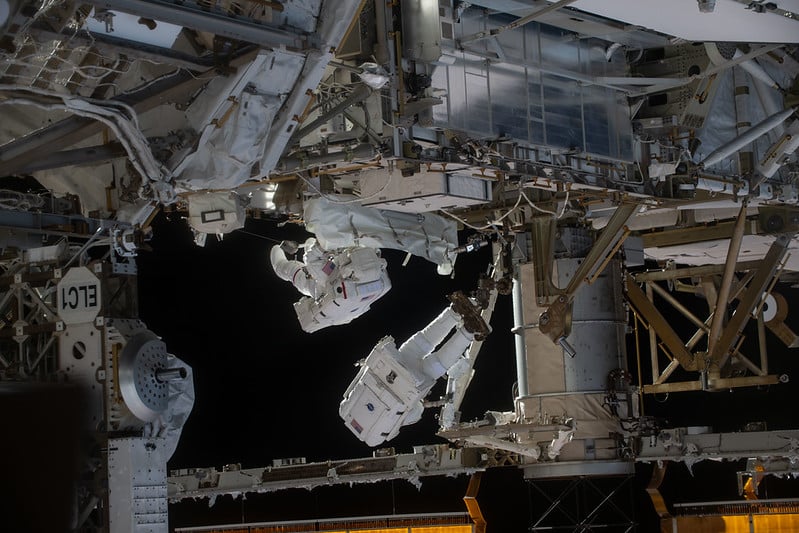 Two astronauts wearing white space suits perform a spacewalk outside the International Space Station, surrounded by metallic structures and equipment, with the blackness of space in the background.