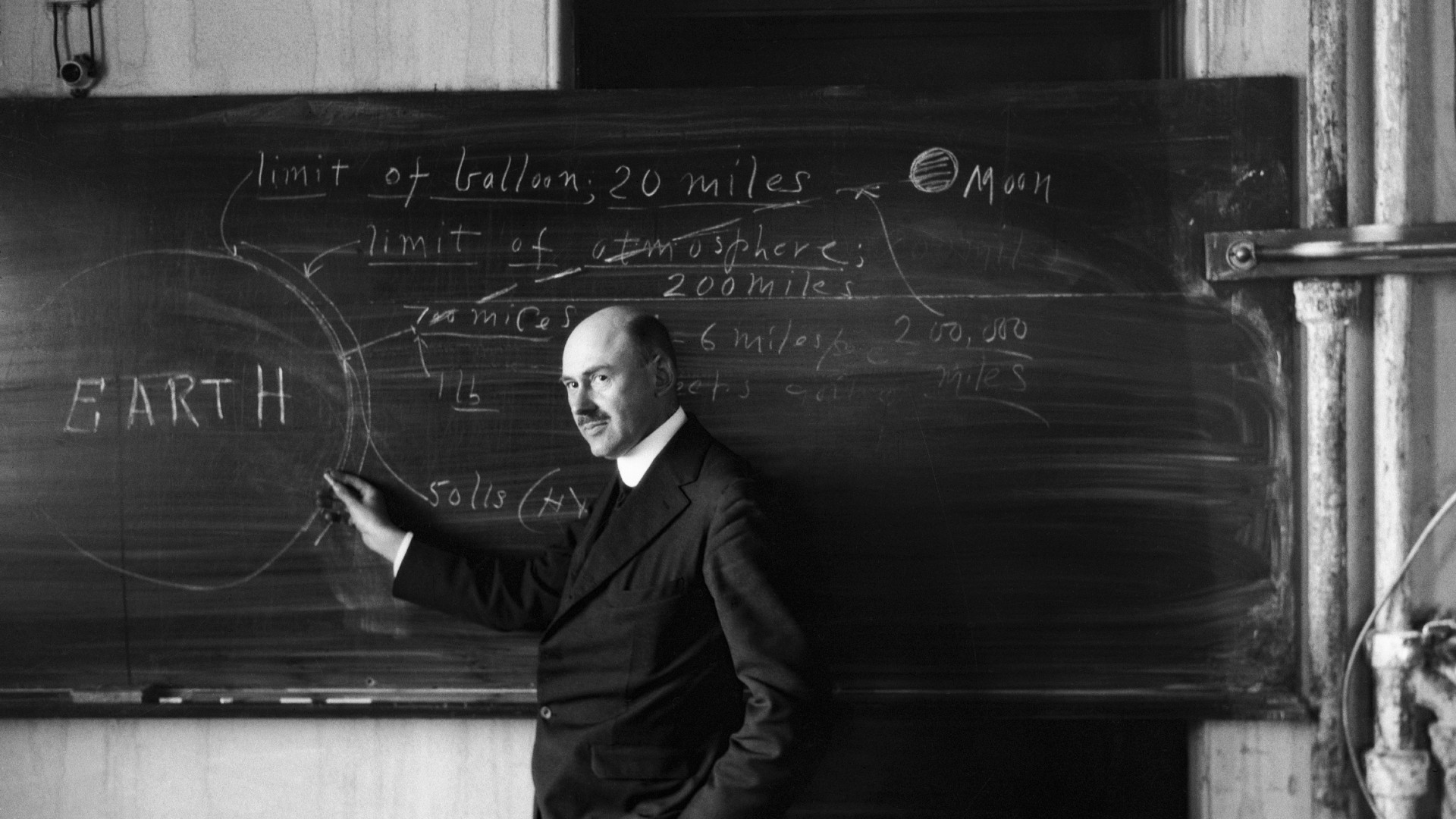a man in a dark suit stands at a chalkboard upon which a large circle labelled "earth" has been drawn, next to a smaller circle labelled "moon"