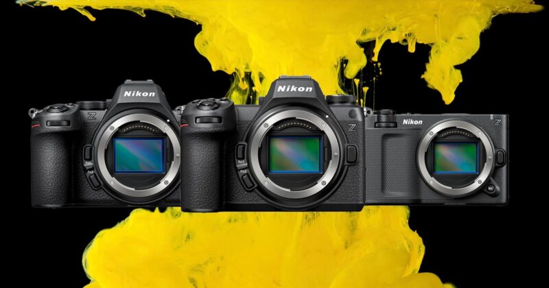 Three Nikon mirrorless cameras are lined up side by side, with their lens mounts exposed, against a background of vibrant yellow paint splashes on black.