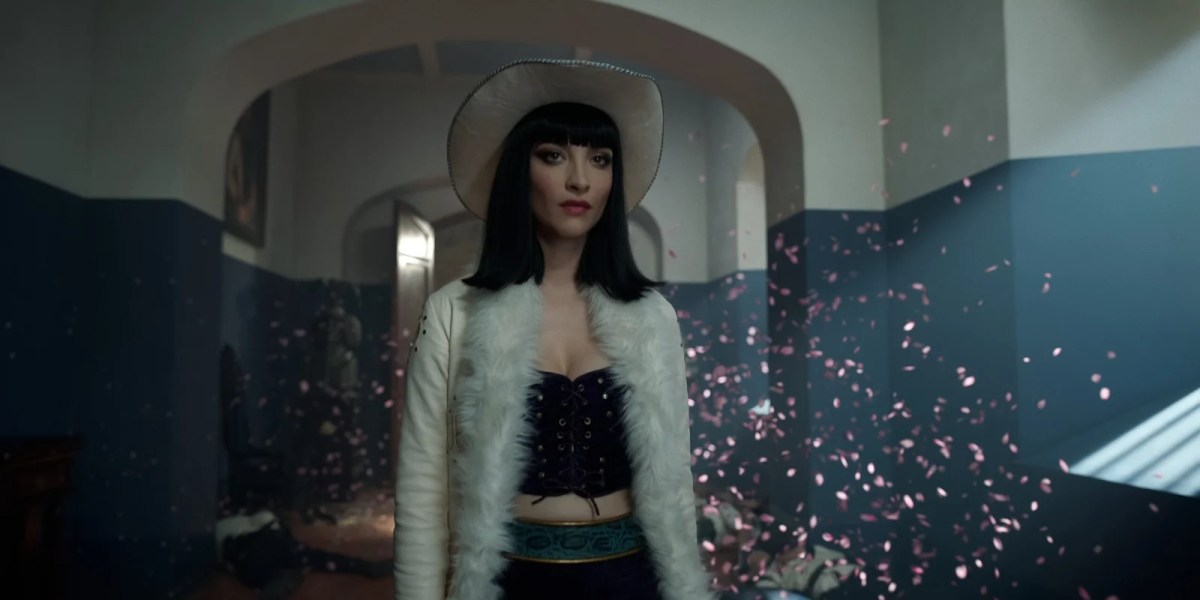 Miss All Sunday standing in a white coat with flower petals falling around her in One Piece Season 2.