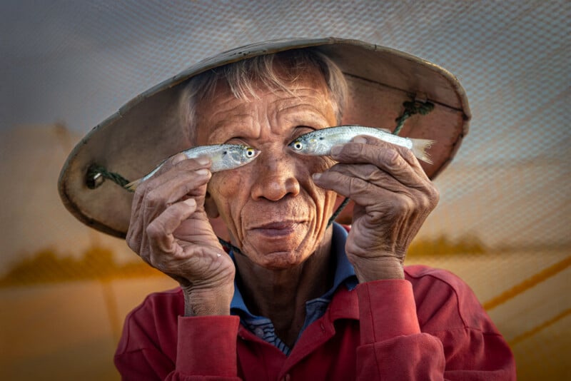 An elderly person wearing a conical hat holds two small fish up to their eyes, creating the illusion of fish eyes. The person is smiling gently and wearing a red shirt, with a net and warm lighting in the background.