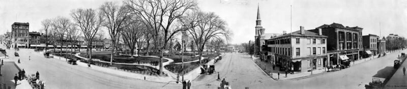A black-and-white panoramic photo of an old town square with leafless trees, surrounding buildings, horse-drawn carriages, and people walking along wide streets. A church steeple is visible on the right.