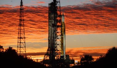 An orange rocket with white side booster stands next to the launch tower during a colorful sunrise.