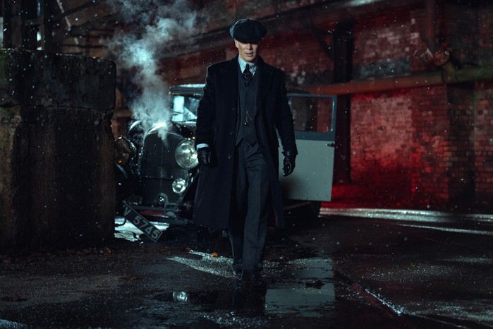 Cillian Murphy in Peaky Blinders: The Immortal Man A man (Cillian Murphy) in a long dark overcoat and flat cap walks down a wet, dimly lit street at night, with snow falling around him and a vintage car steaming behind him near a brick wall illuminated by red light.