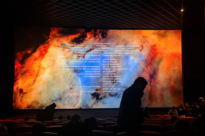 A movie theater screen displays colorful swirling patterns and film credits, while silhouetted audience members are seated and standing in front of the screen. The theater has red seats and a dark ceiling.