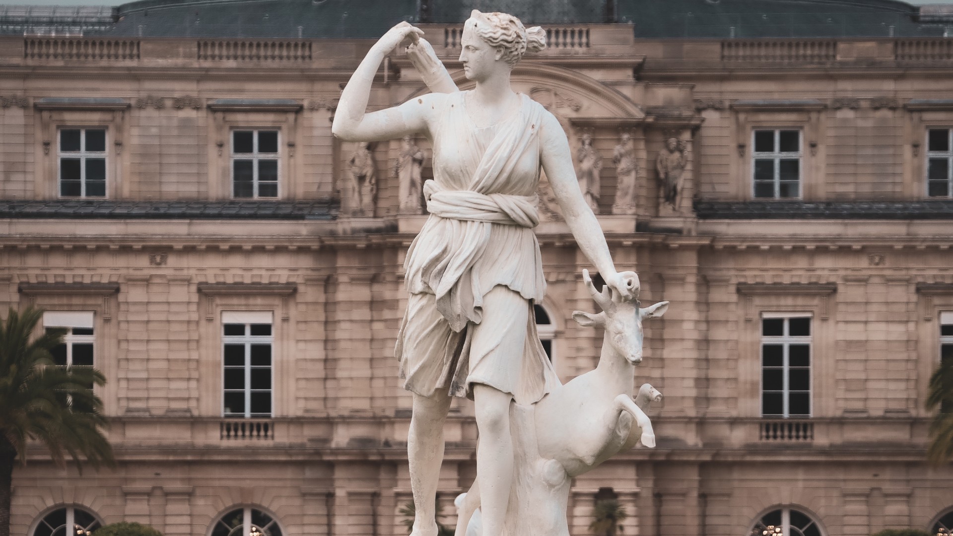 a white statue of a female figure in a toga holding a quiver of arrows, placing her hand on the head of a deer