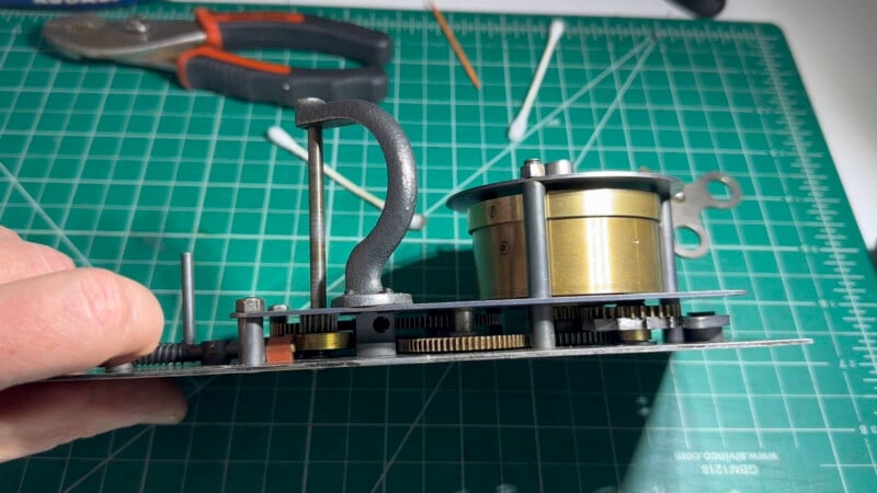 A close-up side view of intricate clockwork gears and mechanisms on a green cutting mat, with pliers, cotton swabs, and a wooden stick visible in the background.