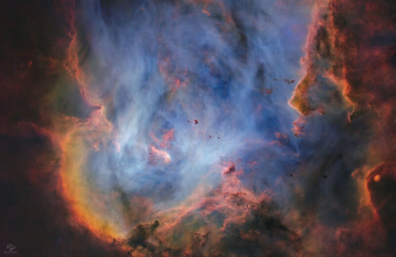 Colorful nebula with swirling clouds of blue, orange, and pink gases; dark silhouettes of gas and dust appear to form the outlines of faces on the right side of the image.