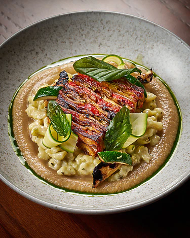 Gourmet plated dish of grilled vegetables on creamy pasta, garnished with fresh herbs and zucchini ribbons.