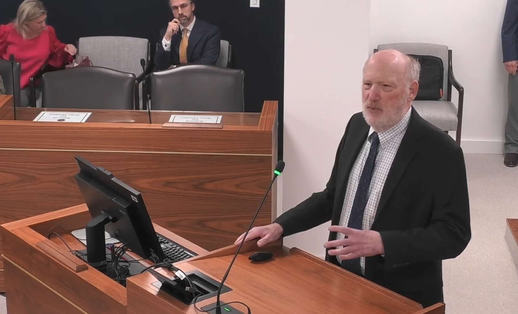 A man in a suit stands at a podium talking about outpatient commitment at a legislative IVC meeting