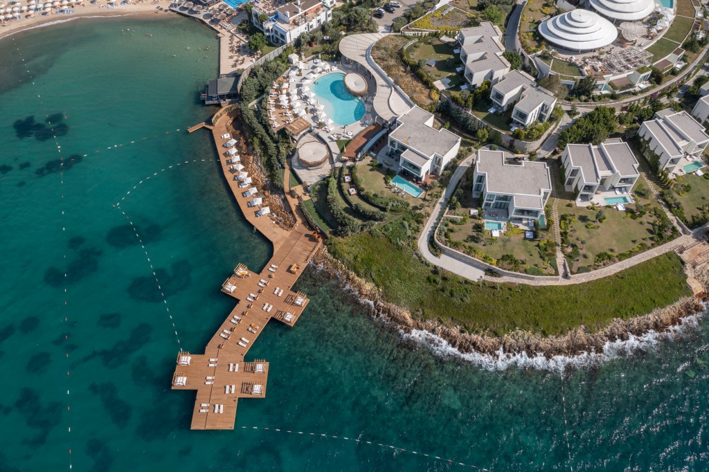 Coastal resort with white buildings, private pools, wooden pier with loungers, circular pool, and rocky shoreline by the sea.