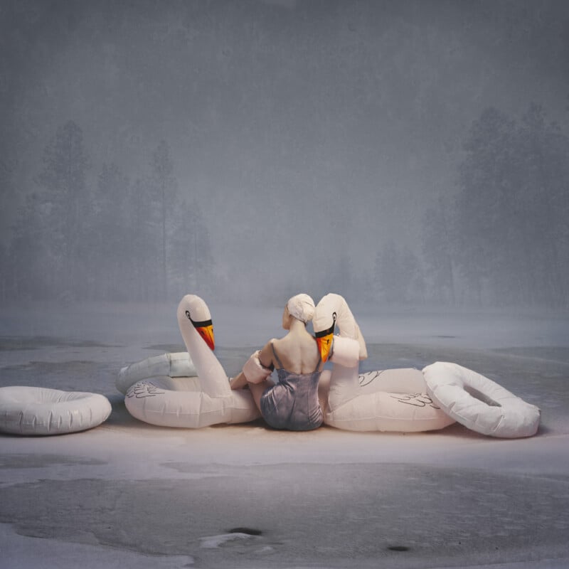 A person in a bathing suit and swim cap sits on an inflatable swan, surrounded by deflated pool floats, on a frozen, snowy landscape with trees in the background.