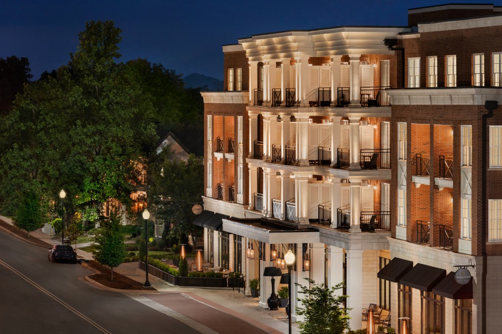 Multi-story brick building with white columns, balconies, and lit windows at night, set along a quiet, tree-lined street.