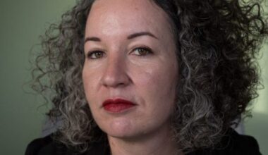 A woman with curly gray hair, wearing a black top and gold necklace looks directly at the camera with a serious expression against a neutral background.