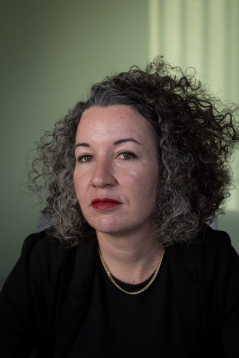 A woman with curly gray hair, wearing a black top and gold necklace looks directly at the camera with a serious expression against a neutral background.