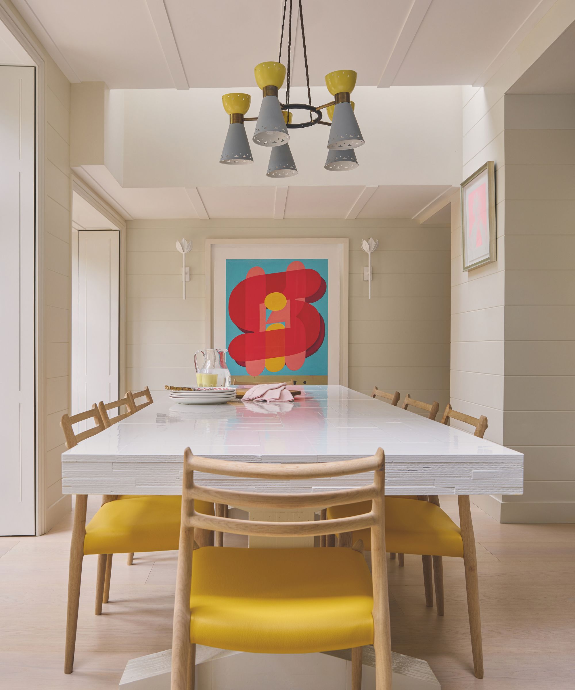 A modern dining room featuring a long, white high-gloss table surrounded by wooden chairs with bright yellow seats. A colorful abstract graphic print hangs on the paneled wall, complemented by a mid-century chandelier with grey and yellow shades.