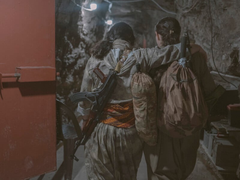 Two people with long hair, dressed in camouflage clothing, walk side by side in a dimly lit underground tunnel. One carries a rifle and both have bags slung over their shoulders, with one arm draped around the other's back.