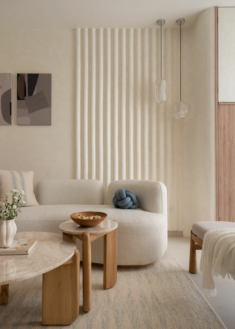 Minimalist living room with cream-colored textured furniture, wooden tables, abstract wall art, pendant lights, and a ribbed accent wall. Neutral and beige tones dominate the decor.