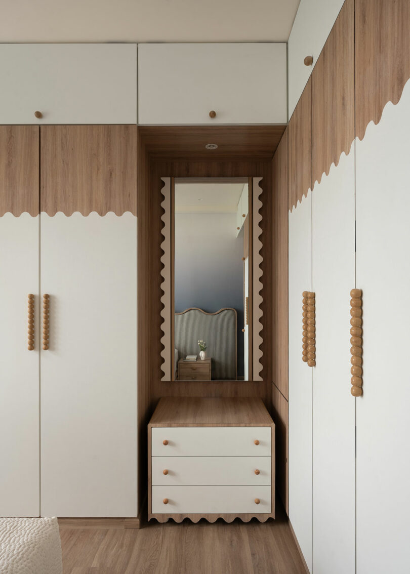 A built-in wardrobe with white doors, wooden trim, scalloped edges, a central mirror, and a white chest of drawers on a wooden floor.