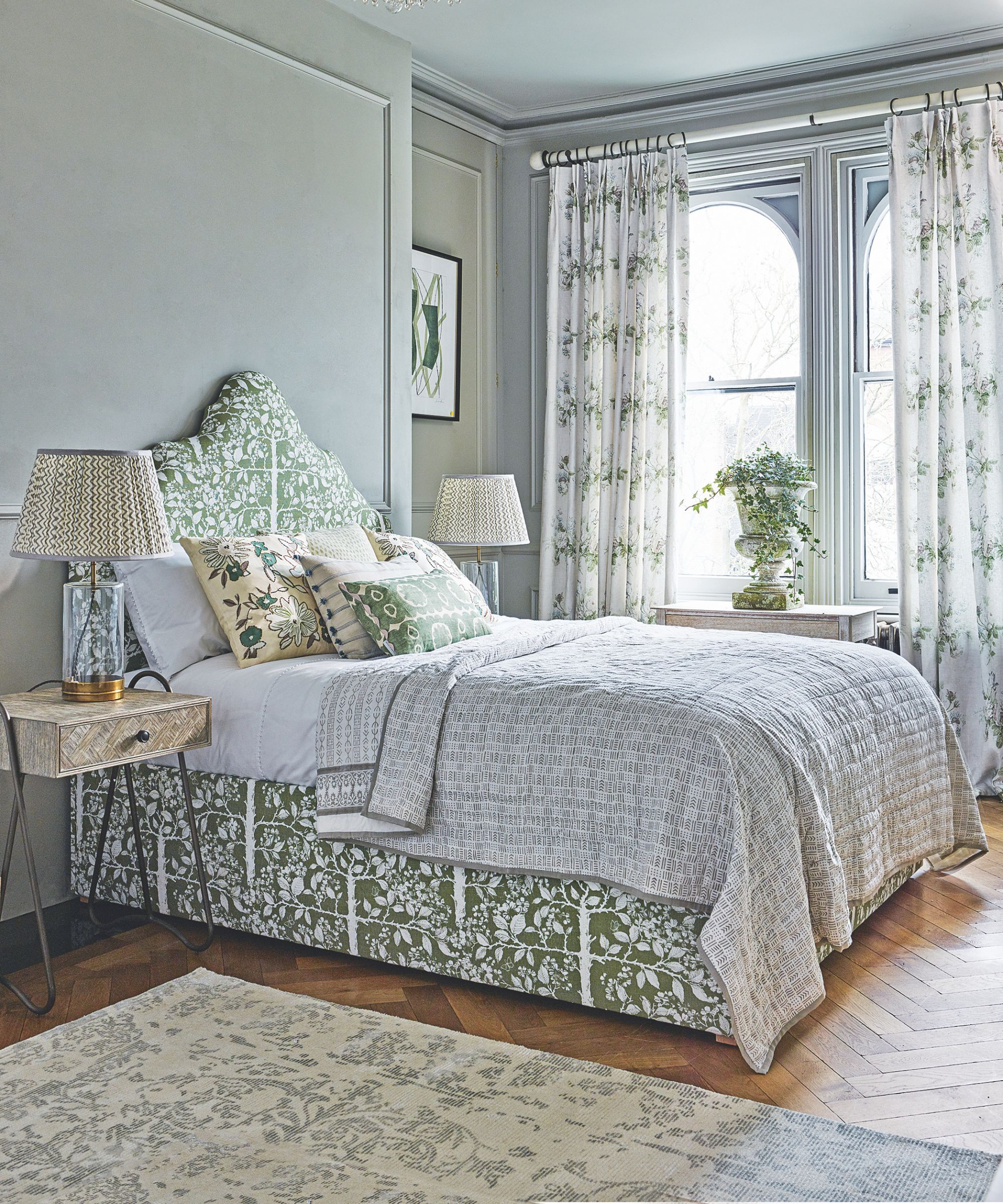 A pale green bedroom space with sage panelled walls, a green patterned double bed with headboard, with patterned bedside tables, and patterned lamps to either side. The floor is light wood and polished, and in the foreground is a large patterned rug. In the background are two bright arched windows with white and green floral-patterned curtains, and a white plant pot on the centre of the windowsill.