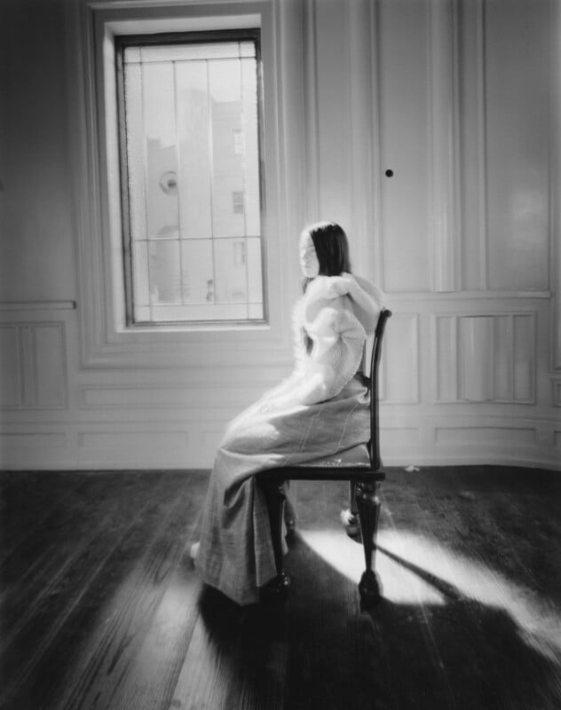 A woman in a long skirt and textured top sits sideways on a chair in a mostly empty room with wood floors and paneled walls, facing a window with sunlight streaming in. The scene appears soft and ethereal in black and white.