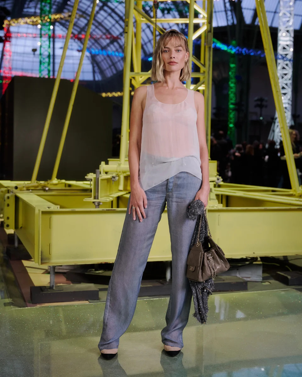 Person on an industrial-style walkway wearing a sheer tank top and wide-leg jeans, holding a purse