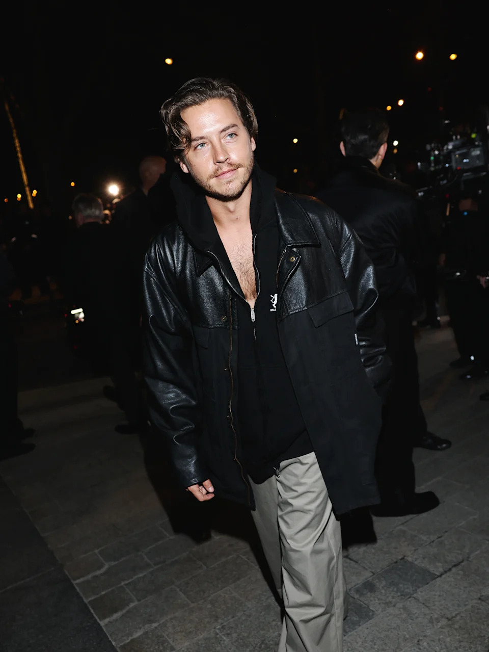 Person in a black leather jacket and a black hoodie at a nighttime outdoor event, walking casually