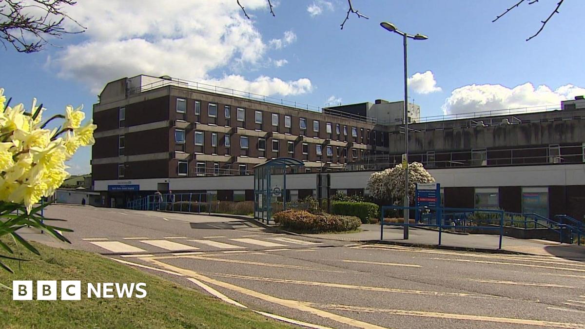 The outside of a hospital on a clear sunny day. The building has lots of windows and has the usual NHS branding on the front of it. There is a road in front of it with some flowers to the left of the pictures.