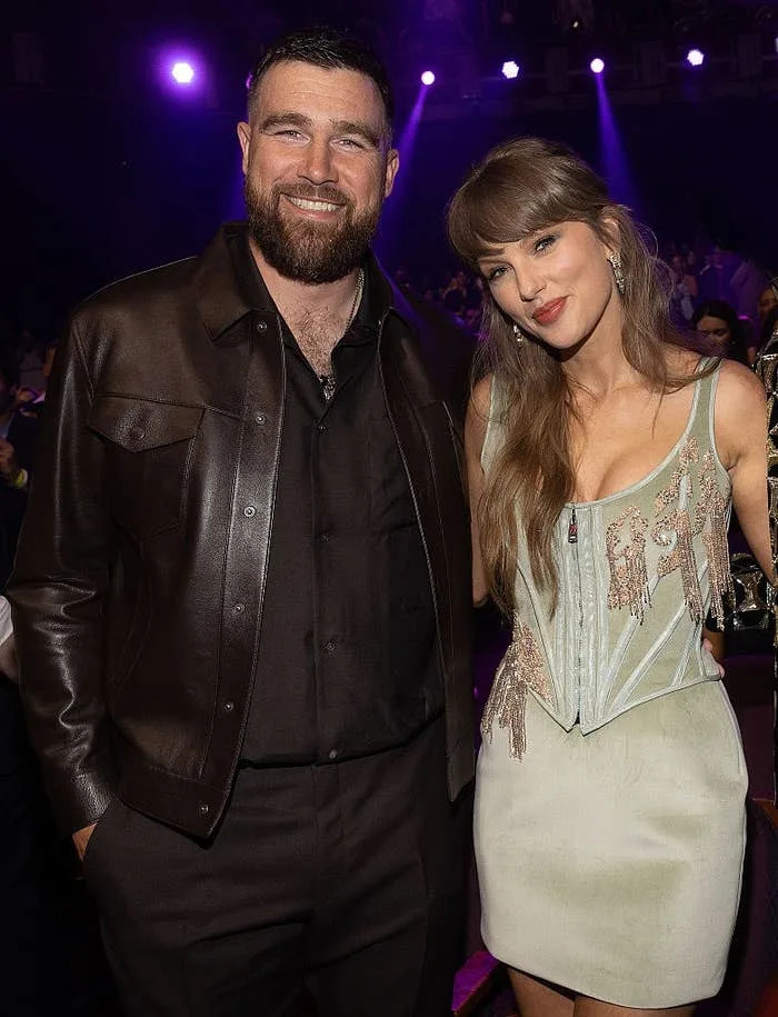Two people pose together at an event. One wears a leather jacket, and the other wears a stylish dress with a corset detail