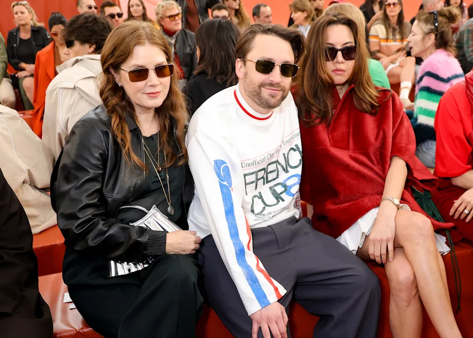 Three people wearing stylish casual clothing with sunglasses sit in a crowded outdoor event setting