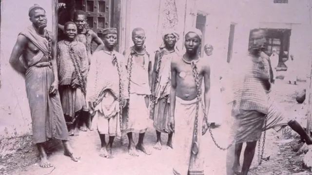 A 19th Century photograph wey show black enslaved pipo in chains for one market on di island of Zanzibar. Dem dey look at di camera.