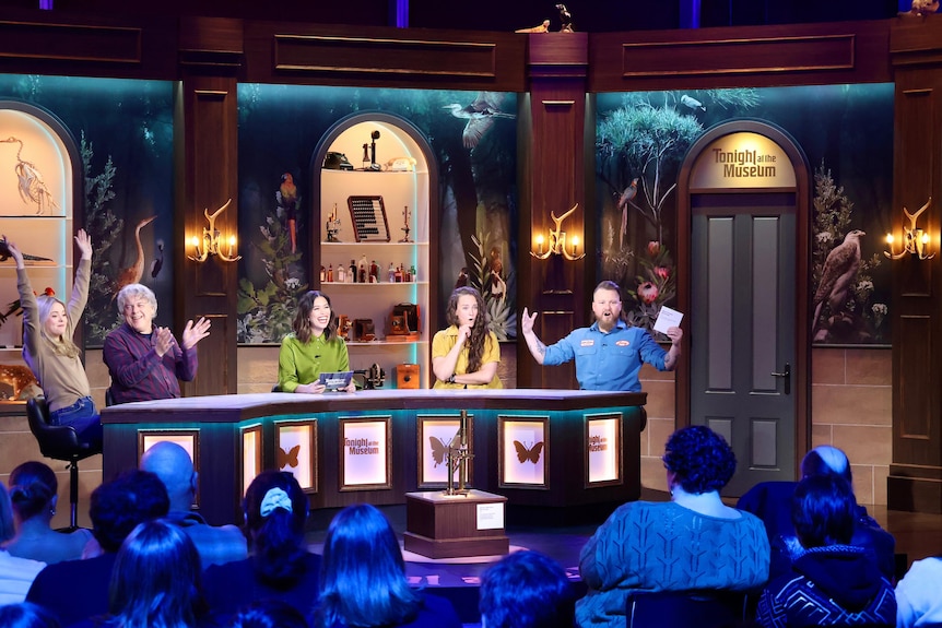 Tegan Higginbotham, Alan Davies, Alex Lee, Zoe Coombs Marr and Brett Blake sit behind a game show table in front of audience.