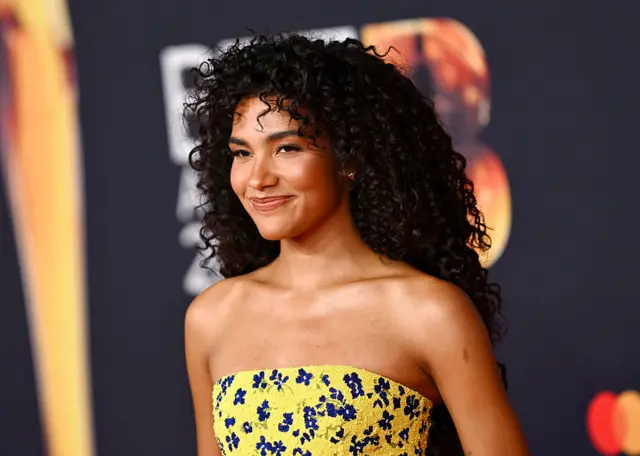 Olivia Dean in a yellow dress with blue flower details, smiling at the camera at the Brit awards.