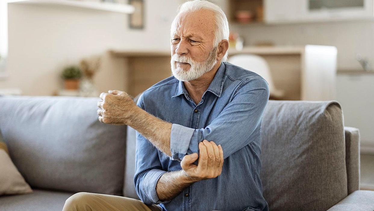 Older man holding elbow in pain while sitting on couch.