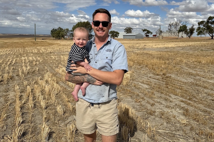 A man in a blue shirt and beige shorts, smiling while wearing sunglasses and holding a baby boy in a grain field.