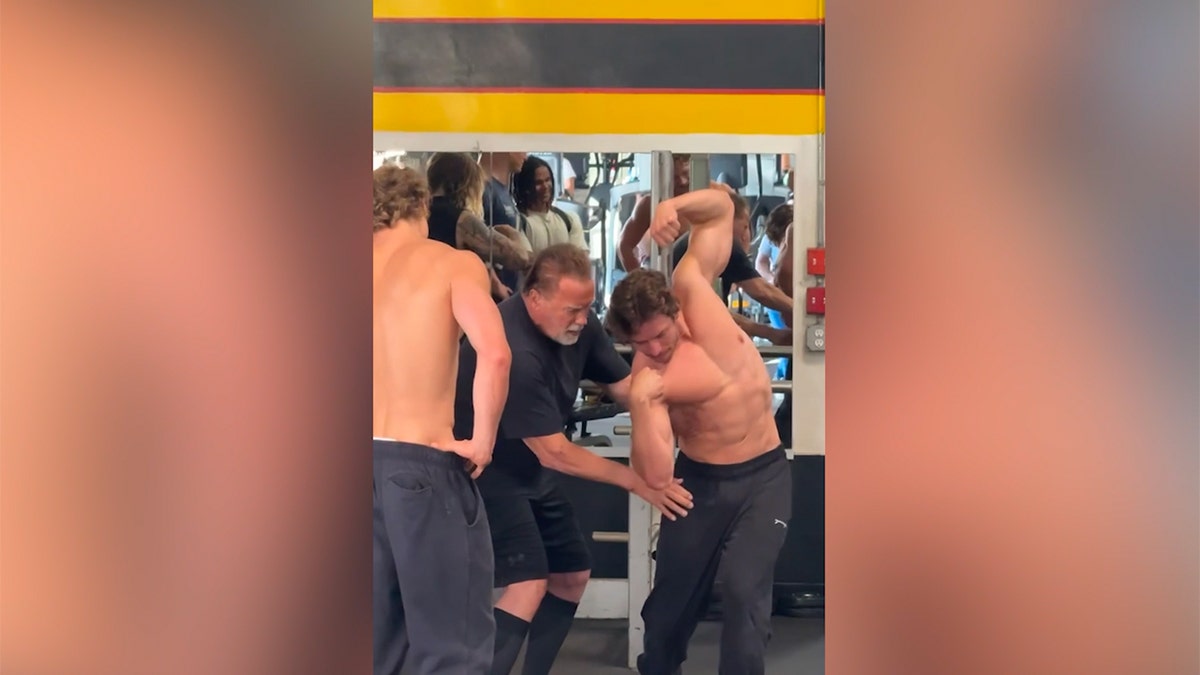 Arnold Schwarzenegger assists Joseph Baena with a bodybuilding pose inside a gym.