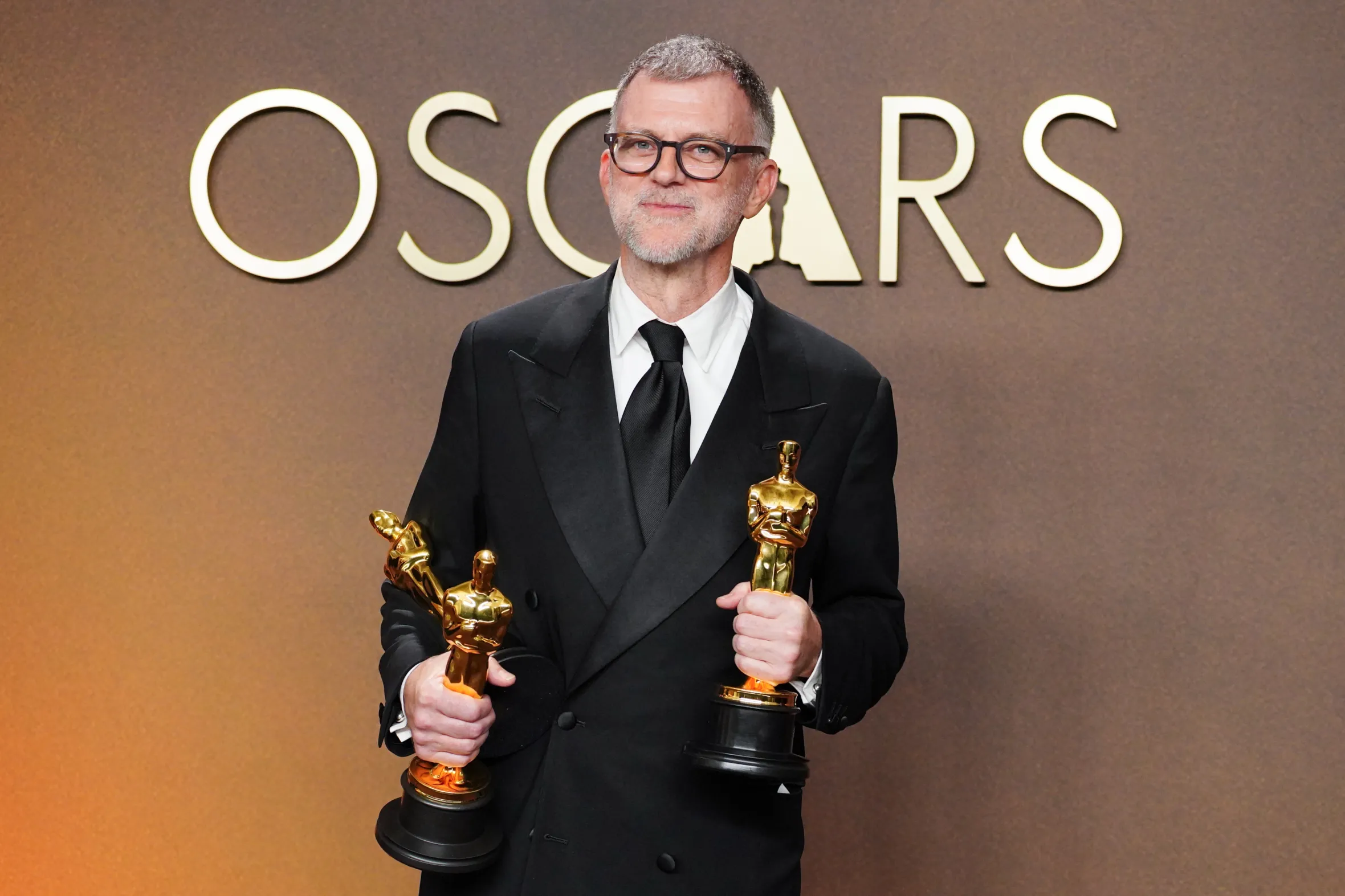 Paul Thomas Anderson holds three Oscar trophies in front of an "OSCARS" sign.