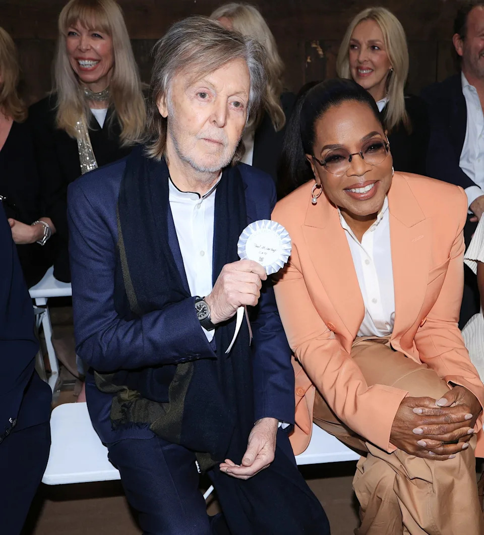 Two public figures seated and smiling at an event, one holding a white ribbon; surrounded by other attendees