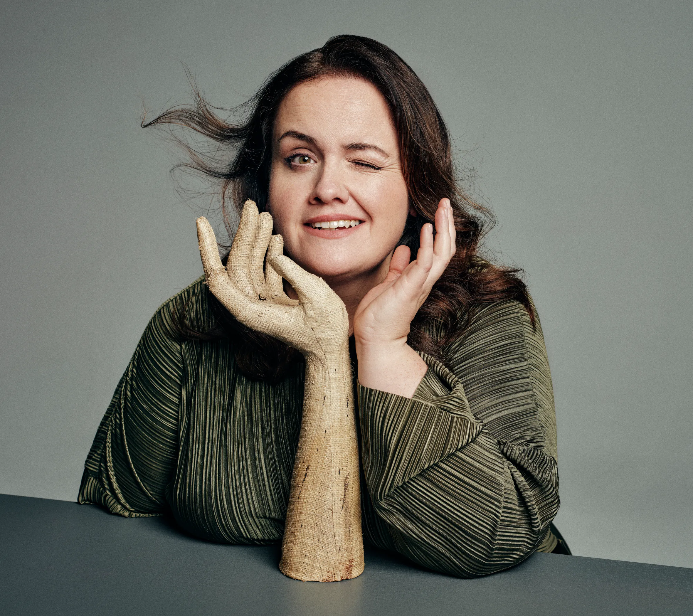 Portrait of Jessica Gunning winking, with a wooden hand prop on the table beside her.