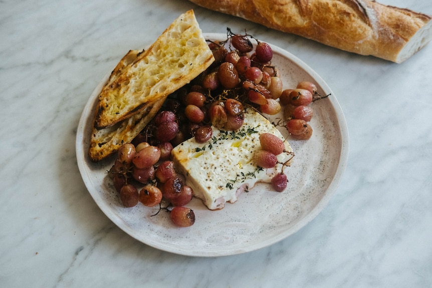 A block of feta cheese and red grapes, roasted on a tray with thyme to create a simple meal when served with a baguette