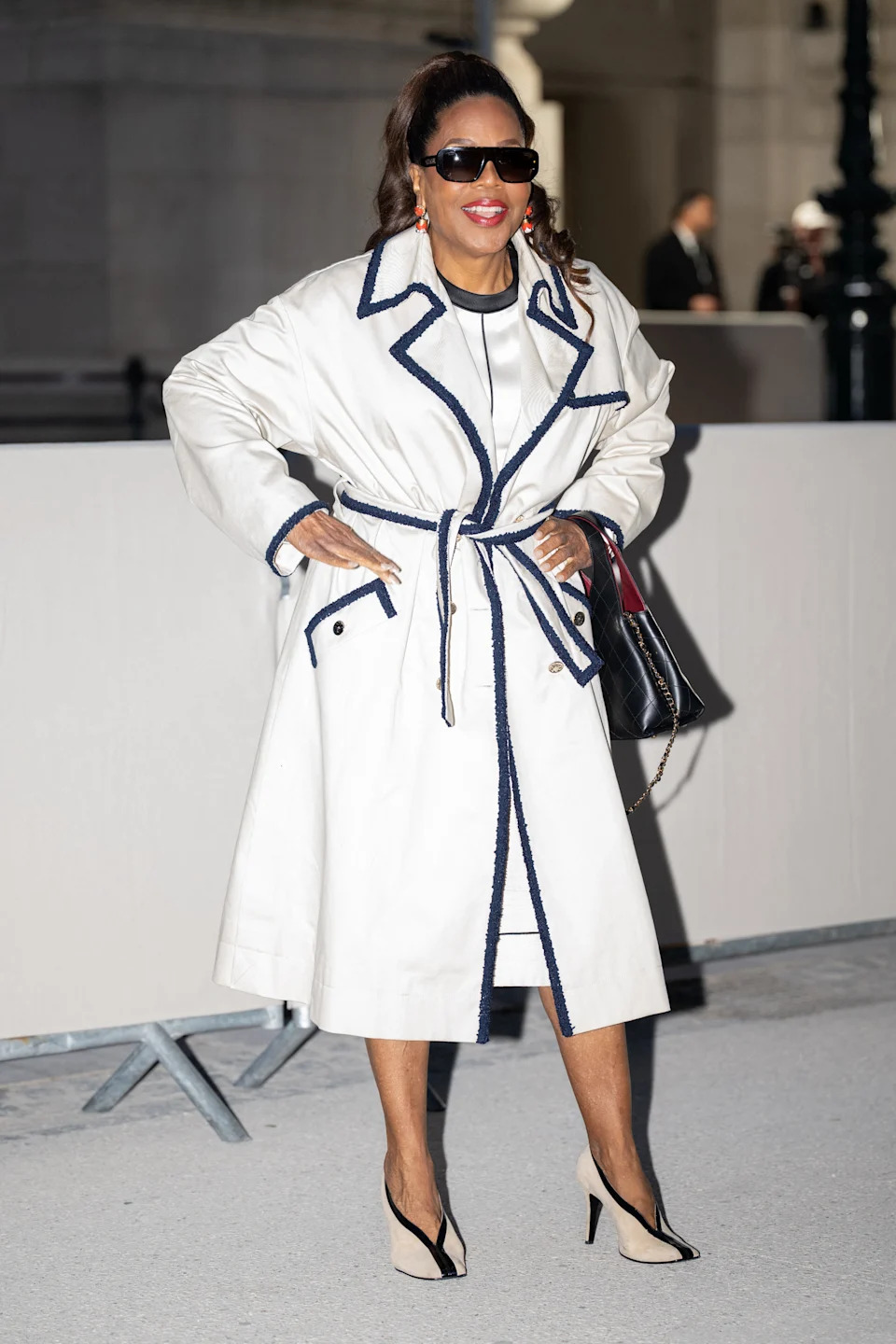 Person in a stylish belted trench coat and sunglasses poses confidently outdoors