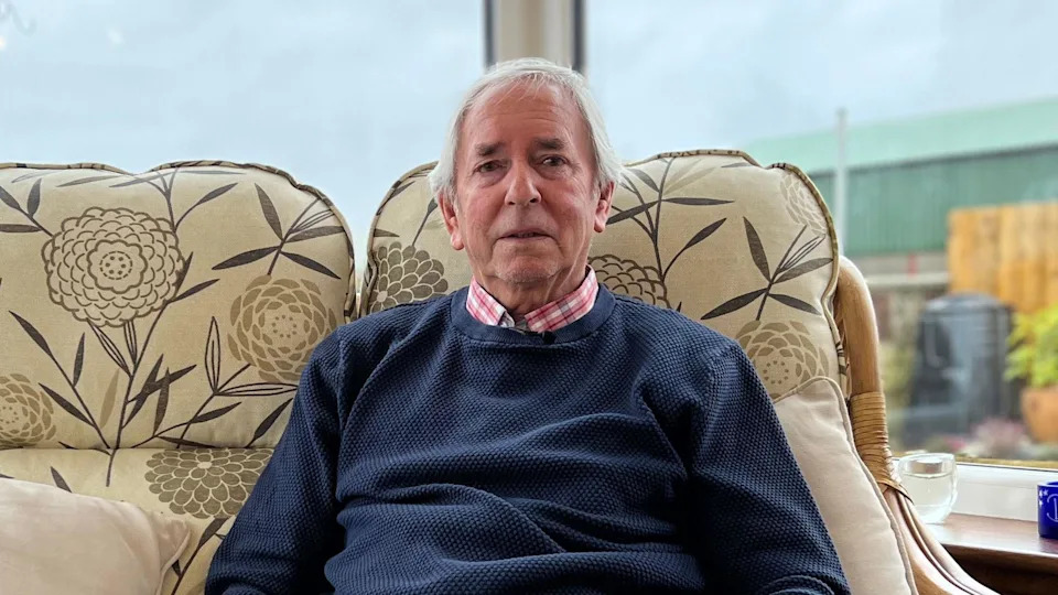 Dave Westren looking into the camera. He has short, white hair and is wearing a blue jumper over a pink and white checked shirt. He is sitting on a beige, patterned sofa with flowers on it. It is in front of a large window which looks out into a garden and there looks to be a barn on the right.