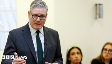 Keir Starmer, in a suit and tie,  speaking while two women in the background watch on