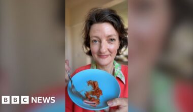 A woman with brown hair holds up a blue plate holding kimchi. She is wearing a dark pink jumper and a green-collared shirt.
