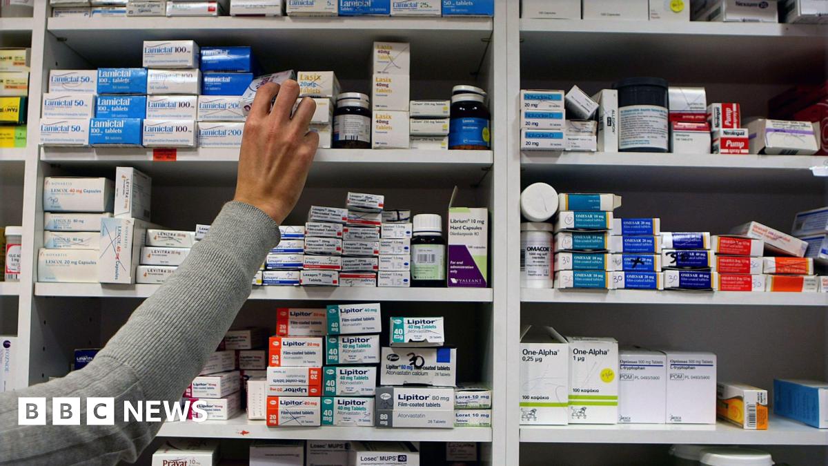 An arm with a grey sleeve is reaching out to a set og shelves packed with a variety of medications