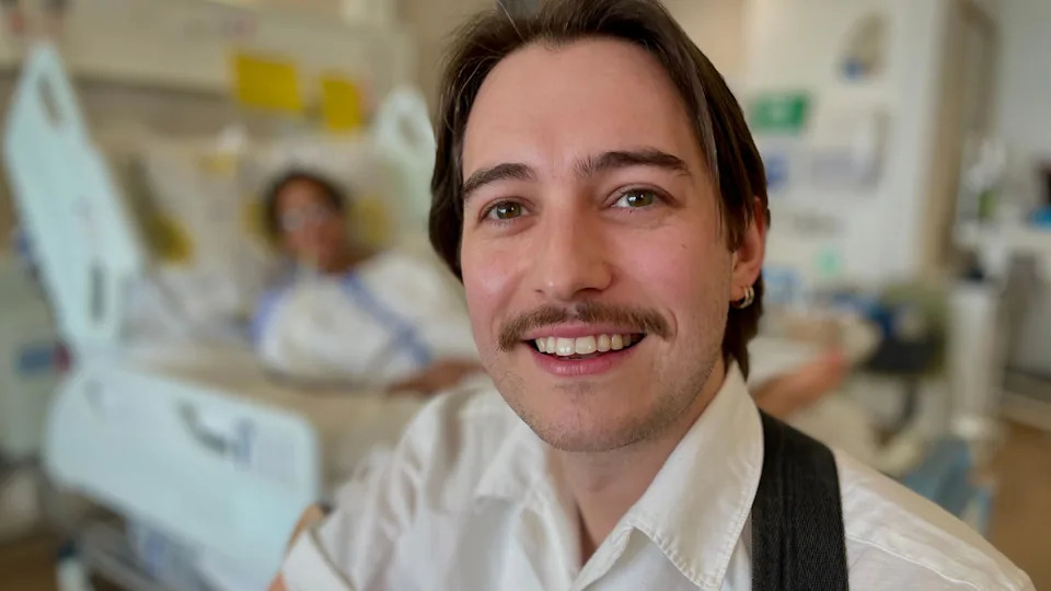Music therapist Joe Jezard smiles at the camera sitting in a hospital ward with one of his therapy patients in the background 