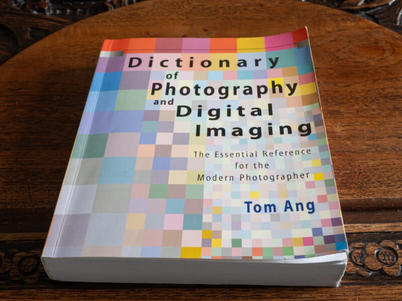 A book titled "Dictionary of Photography and Digital Imaging" by Tom Ang lies on a wooden surface. The cover features a colorful mosaic pattern and subtitles it as "The Essential Reference for the Modern Photographer.