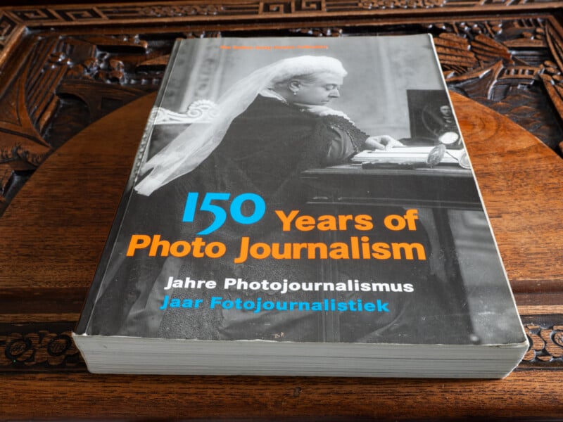 A book titled "150 Years of Photo Journalism" rests on a carved wooden surface. The cover features a black and white photograph of an elderly woman in formal attire, with text in English, German, and Dutch.